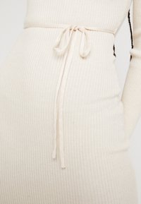 Geripptes cremefarbenes Kleid mit einer figurschmeichelnden Silhouette und einem passenden Stoffgürtel, der in der Taille gebunden ist. Die Textur ist weich und dehnbar.