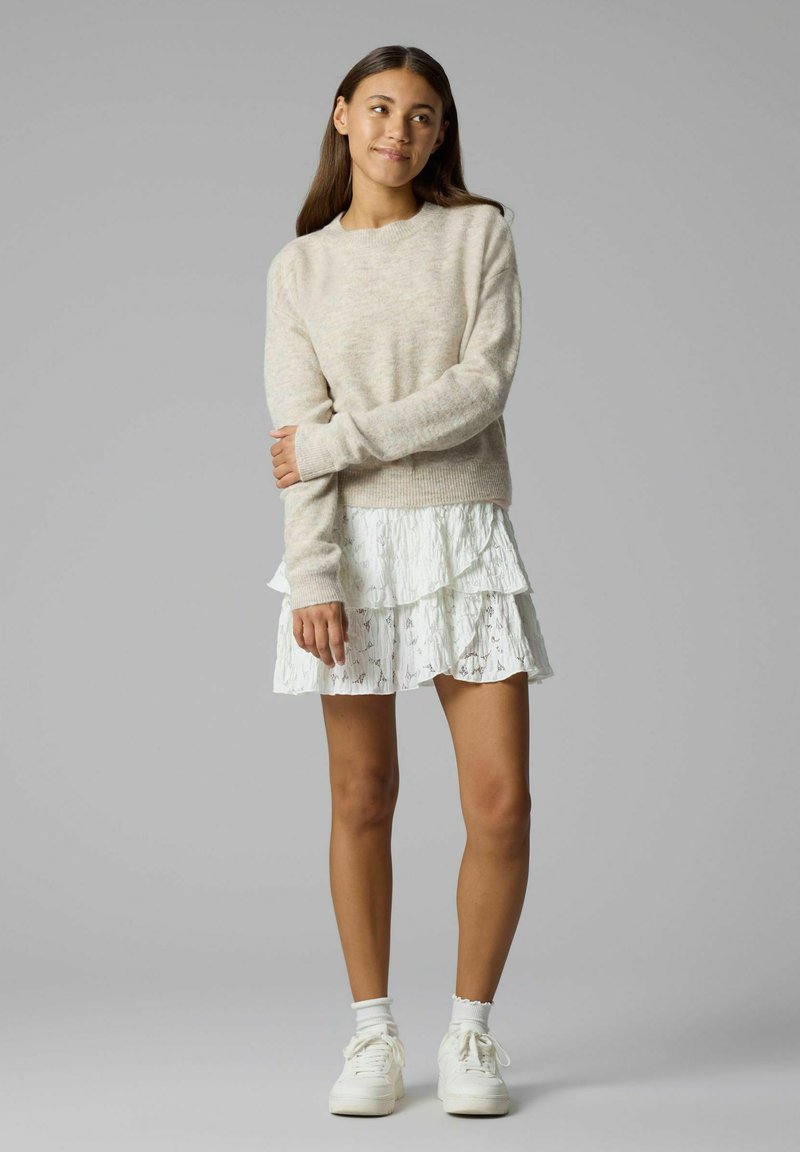 Jeune femme aux longs cheveux bruns portant un pull beige, une jupe blanche superposée, des chaussettes blanches et des baskets blanches, debout, les bras croisés.