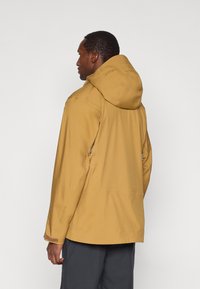 Żółta kurtka z kapturem o gładkiej fakturze i regulowanych elementach; widoczne szwy i zamki; noszona na ciemnych spodniach.