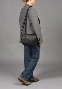 Bolso cruzado negro tejido con un patrón texturizado, que cuenta con una correa larga y un cierre de cremallera, llevado junto a un blazer gris y jeans.