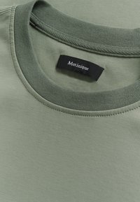 T-shirt vert clair avec une texture lisse, présentant un col rond et une étiquette sombre portant l'inscription "Matinique" cousue dans le tissu.