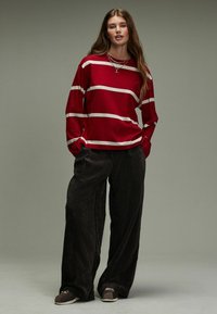 Rotes Langarmshirt mit weißen horizontalen Streifen, kombiniert mit dunklen, strukturierten weit geschnittenen Hosen und lässigen braunen Schuhen.