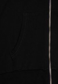 Svart zip-up hettegenser med frontlomme, ribbet nederkant og kontrasterende sømdetaljer langs glidelåsen. Myk, teksturert stoff.