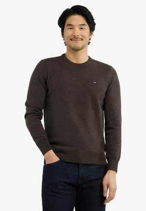 Homme aux cheveux foncés sourit, portant un pull marron foncé ajusté et un jean bleu foncé, debout avec une main dans la poche.