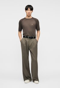 Homme portant une chemise marron à manches courtes, un pantalon taupe à jambes larges avec une ceinture noire, des baskets blanches, debout les mains dans les poches devant un fond uni.