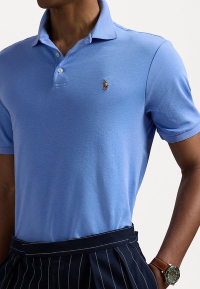 Camisa polo azul claro de suave tejido, con cuello, tres botones y un pequeño logotipo bordado. Combinada con pantalones marinos con pliegues.
