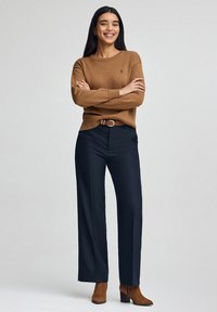 Brązowy dziergany sweter z okrągłym dekoltem i detalem z logo; zestawiony z granatowymi spodniami o szerokich nogawkach oraz beżowymi zamszowymi botkami na kostkę.