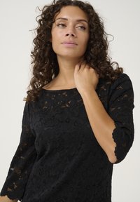 Black lace top featuring a floral pattern, round neckline, and three-quarter sleeves with scalloped edges. Soft texture and semi-transparent design.