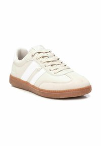 Zapatilla deportiva beige con parte superior de ante texturizado, diseño de rayas blancas, suela de goma marrón clara y cordones planos. Punta redondeada y cuello acolchado.