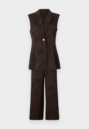 Γυναίκα χαμογελά, στέκεται με τα χέρια σταυρωμένα, φοράει αμάνικο σκούρο σακάκι με σχέδια, παντελόνια με φαρδιά πόδια και ψηλοτάκουνα ανοιχτά παπούτσια, σε λευκό φόντο.