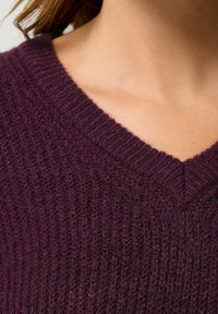 Sweter w kształcie litery V w głębokim fioletowym kolorze, z teksturowanym wzorem dzianiny i ribbowymi wykończeniami. Materiał wydaje się miękki i gruby, odpowiedni do zapewnienia ciepła.