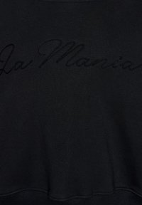 Czarna tkanina z delikatnym, wypukłym, haftowanym kursywą napisem "La Mamania" na środku.