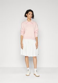 Suéter polo de manga larga en rosa claro, con cuello, combinado con una falda blanca plisada y zapatillas blancas. Diseño sencillo, ajuste relajado.