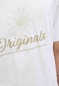 Camiseta de algodón blanca con un gráfico de estrella verde claro y texto dorado "Originales" con detalles de diseño adicionales debajo.