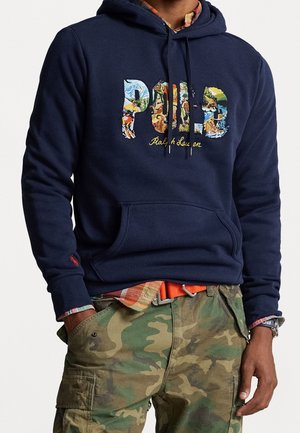 Mand iført marineblå hættetrøje med farverigt Polo Ralph Lauren-logo og grønne camouflage cargobukser med hænderne i lommerne.