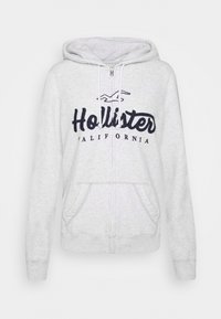 Světle šedá zipová mikina s přední kapsou, vyšitým logem "Hollister" v námořnické barvě a kapucí s nastavitelnými šňůrkami. Měkká textura.