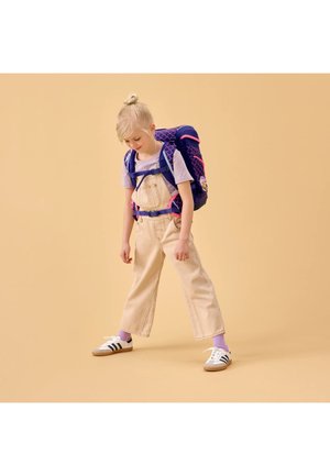 Junges blondes Kind mit hellen Latzhosen und einem lila Hemd, trägt einen großen blauen und pinken Rucksack, steht vor einem beigen Hintergrund.