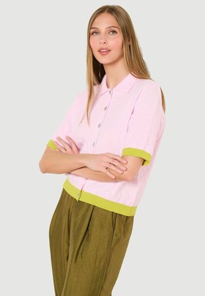 Mladá žena s dlhými blond vlasmi, oblečená v bledoružovom kardigane s krátkym rukávom a žltým lemovaním a olivovo zelených nohaviciach, s prekríženými rukami.