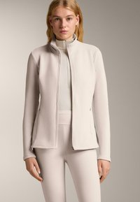 Žena v světle béžové zipové bundě přes odpovídající legíny s vysokým pasem a dlouhým rukávem, stojící proti jednoduchému neutrálnímu pozadí.