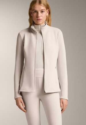 Nő világos bézs színű, cipzáros dzsekit visel, amely alatt illeszkedő, magas derékú leggings és hosszú ujjú felső található, egy egyszerű, semleges háttér előtt áll.