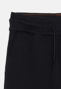 Κοντινό πλάνο της μέσης μαύρου παντελονιού φόρμας με κορδόνι και πλαϊνή τσέπη, με πορτοκαλί ραφές λεπτομέρειας στο εσωτερικό της μέσης.