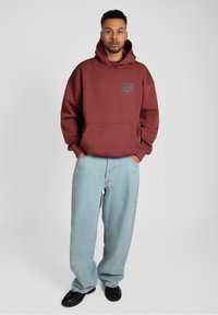 Bordová mikina s predným vreckom a logom; nosená s svetlomodrými voľnými džínsami a čiernymi topánkami. Jednoduché pozadie, uvoľnený strih.