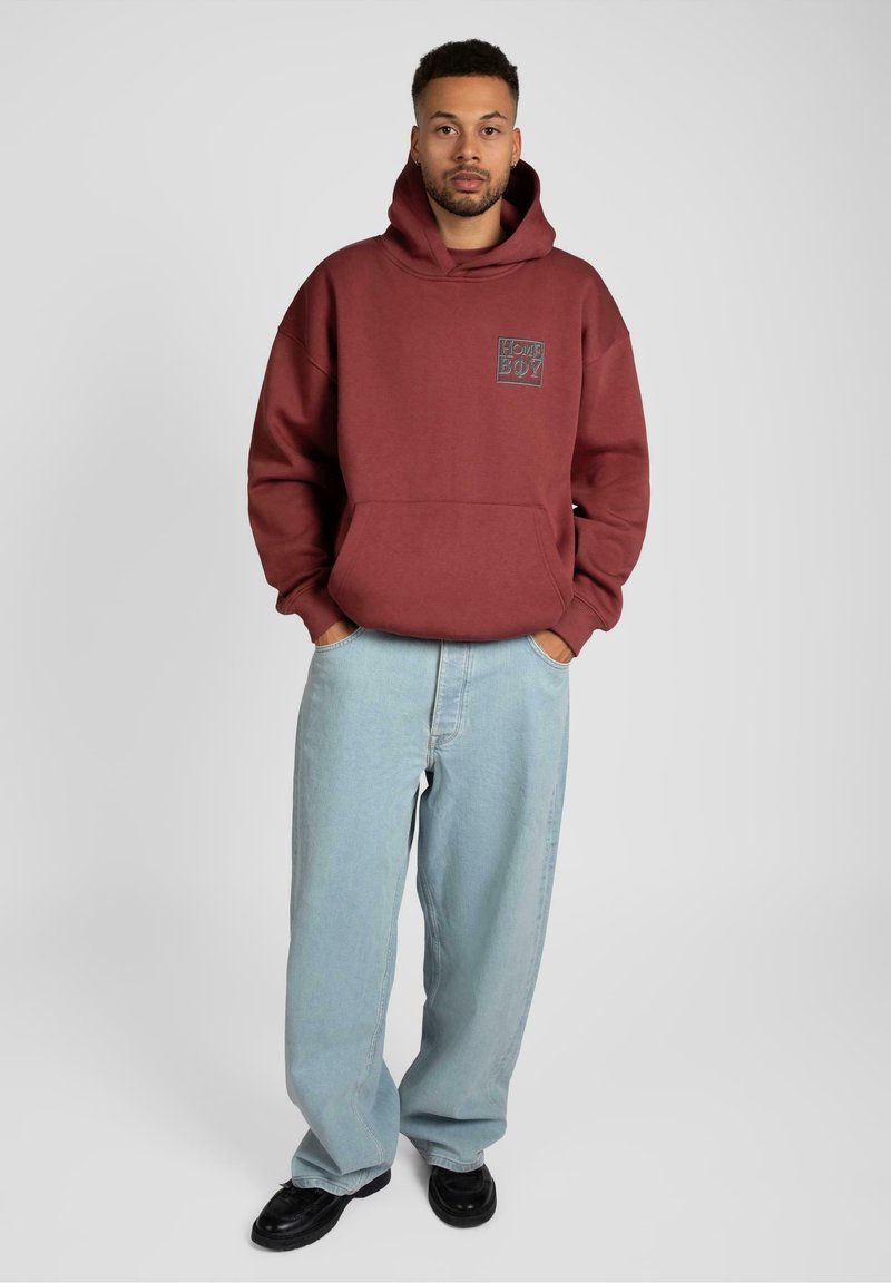 Bordeaux huvtröja med framficka och logo; bärs med ljusblå lösa jeans och svarta skor. Enfärgad bakgrund, avslappnad passform.