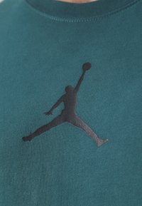 Tealfärgad t-shirt med en svart silhuett av en hoppande figur. Tyget är mjukt, med rund halsringning och korta ärmar.