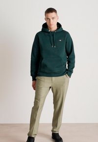Dark green hoodie with a kangaroo pocket and drawstring hood, paired with light grey-green trousers and black sneakers on a neutral background.