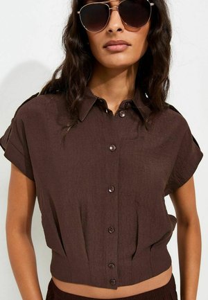 Femme portant un crop top marron foncé à manches courtes boutonné et de grandes lunettes de soleil foncées avec des cheveux ondulés mi-longs.