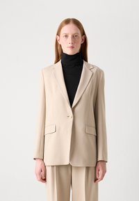 Beige tailored blazer with single button closure, notch lapels, two front pockets, and smooth texture. Worn over a black turtleneck.