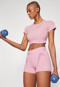 Lys rosa trenings crop top og shorts sett med ribbede detaljer og lilla striper. Holder en blå 3 kg hantel. Jevn tekstur.