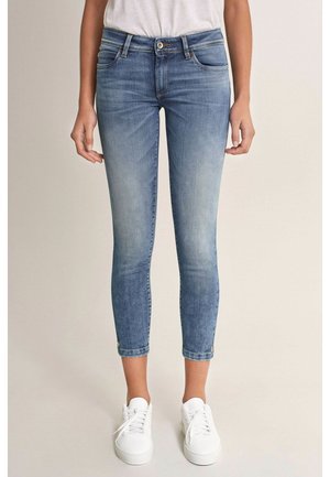 Femme portant un jean skinny cropped bleu délavé à taille moyenne et des baskets blanches, debout devant un fond neutre.