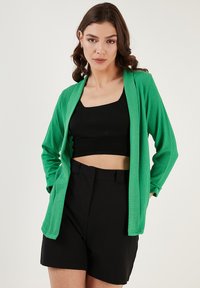 Zelený textúrovaný kardigán s dlhými rukávmi a otvoreným predným dizajnom, nosený cez čierny crop top a čierne šortky.