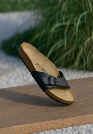 Pojedyncza czarna sandałka Birkenstock z regulowanym paskiem na drewnianej powierzchni, zielone rośliny i biały żwir w tle rozmytym.