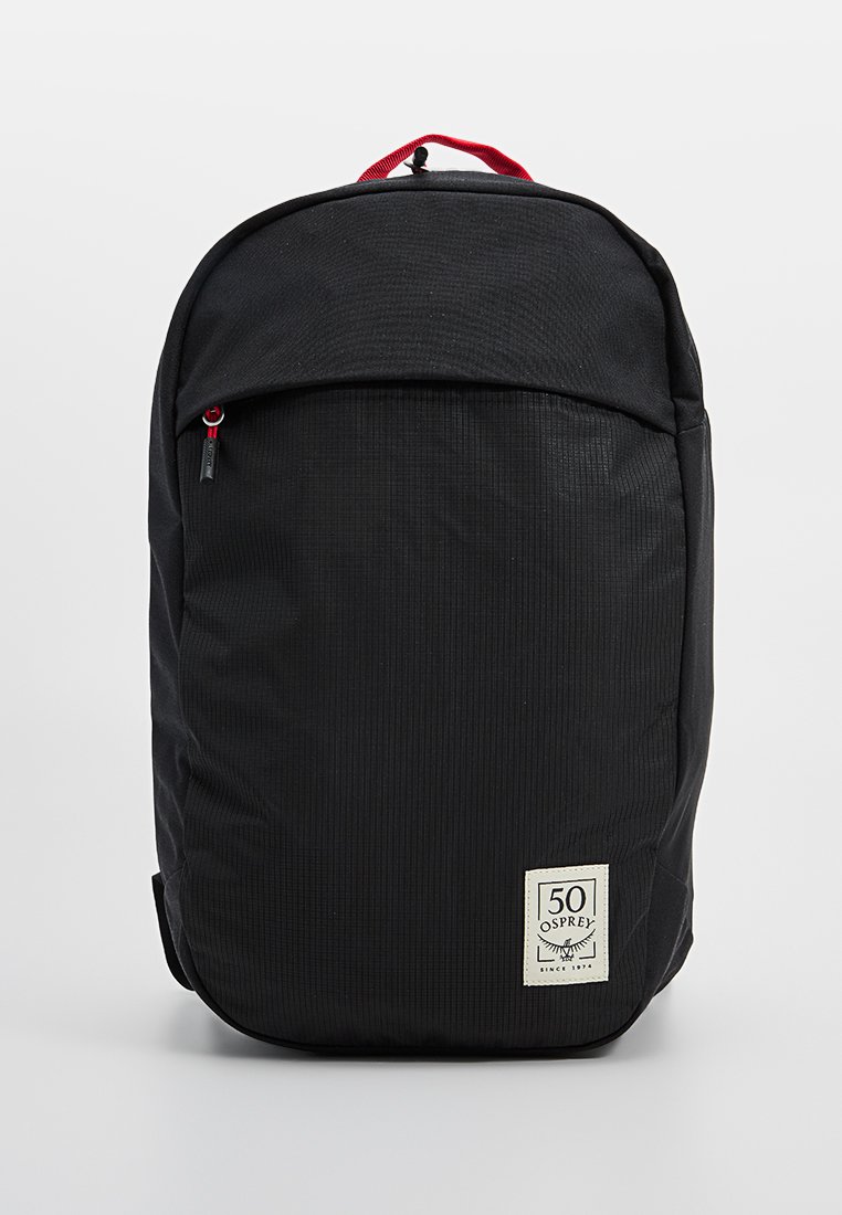 Mochila negra con un tejido texturizado, cierre superior con cremallera, bolsillo frontal y acentos rojos. Presenta un parche de logo en la parte inferior frontal.