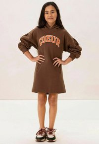 Robe hoodie marron avec des manches oversize, arborant un texte orange "COEUR" et deux poches avant, assortie à des baskets multicolores.