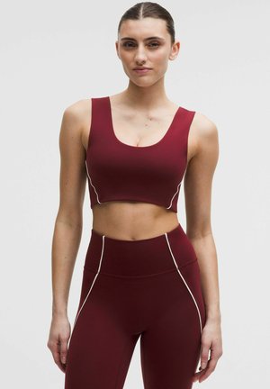 Vrouw die een nauwsluitende bordeauxrode sportbeha en hooggetailleerde legging met witte naadaccenten draagt, staand tegen een effen lichte achtergrond.