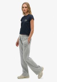 Jonge vrouw staat met haar handen in haar zakken, draagt een marineblauw logo t-shirt, grijze joggingsbroek en beige sneakers op een witte achtergrond.