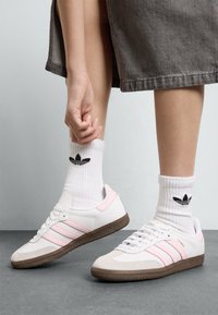 Sapatilhas de couro branco com riscas cor-de-rosa e sola de borracha castanha, combinadas com meias brancas caneladas com um logo preto.