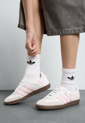 Hvite skinnsneakers med rosa striper og brun gummisåle, sammen med hvite ribbede sokker med svart logo.