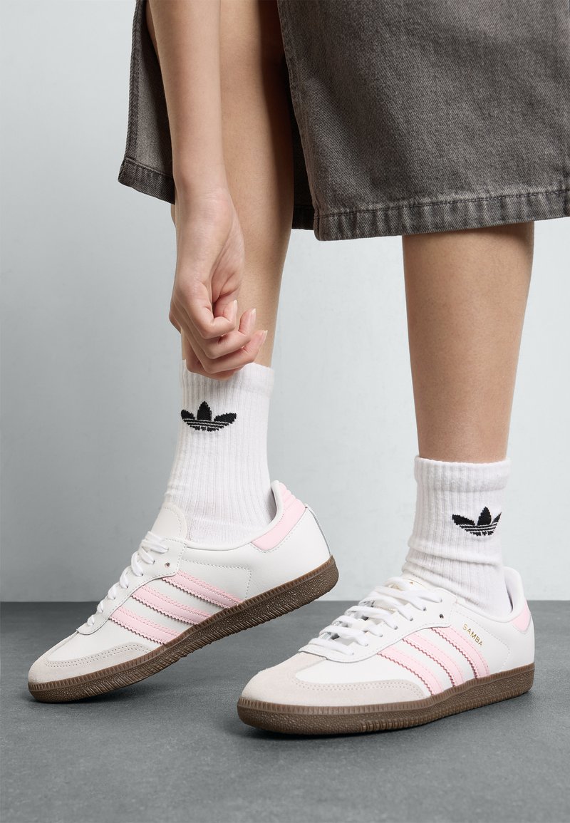 Sapatilhas de couro branco com riscas cor-de-rosa e sola de borracha castanha, combinadas com meias brancas caneladas com um logo preto.