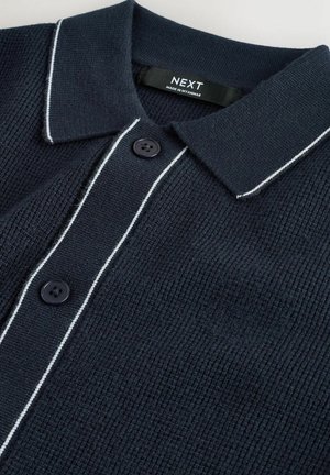 Tamsiai mėlynas megztas polo marškinėlis su tekstūruota medžiaga, baltomis siūlėmis ant apykaklės ir dviem juodais mygtukais ties iškirpte. Matomas prekės ženklo ženklas.