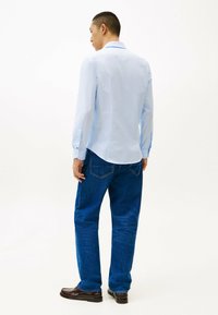 Lichtblauwe blouse met lange mouwen en een rugpas, gecombineerd met blauwe denim jeans en bruine schoenen. Eenvoudige knoopdetails op de manchetten.