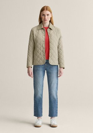 Kvinde iført beige quiltet jakke over rød sweater, blå jeans og hvide sneakers, stående mod en ensfarvet baggrund.
