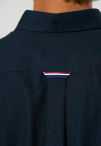 Temno modra srajca z gumbasto ovratnikom, ki ima majhen trobojni poudarek (rdeča, bela, modra) na hrbtni strani blizu ramenskega šiva. Teksturovano blago.