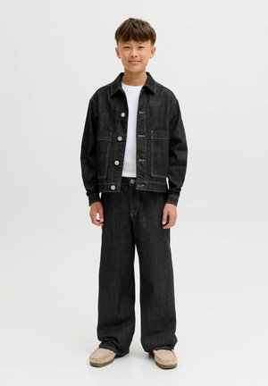 Chlapec stojaci, oblečený v čiernej rifľovej bunde a nohaviciach s bielou košeľou a béžovými mokasínmi na jednoduchom svetlom pozadí.