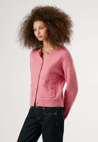 Een model poseert in een roze cardigan met zakken, gecombineerd met donkere jeans. De achtergrond is eenvoudig, waardoor de kleding benadrukt wordt.
