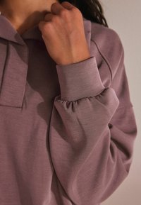 Gara piedurkne puloveri muted mauve krāsā ar brīvu piegriezumu, ar plīšu aproci un smalku tekstūru audumā.