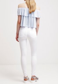 Ljusblå av-axeln-topp med volanglager, matchad med åtsittande vita jeans och silverfärgade sandaler, sedd från baksidan.